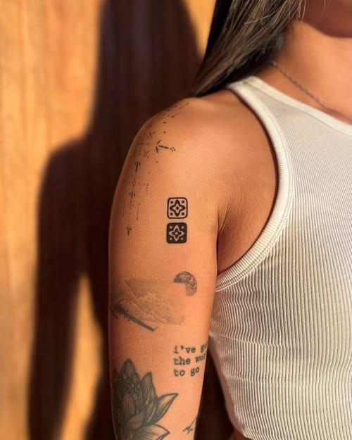 A minimalist black and grey tattoo featuring geometric symbols and script on the upper arm — by Dylan Aiello, los angeles