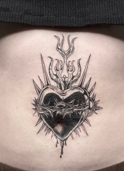 A black and grey tattoo of a heart with thorns and flames on the stomach, featuring bold linework and intricate details — by Shelby Bianco, los angeles
