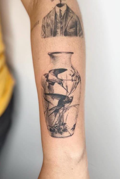 A black and grey realistic tattoo of a bird inside a vase on the forearm, featuring detailed linework and shading — by Level tattoo, los angeles