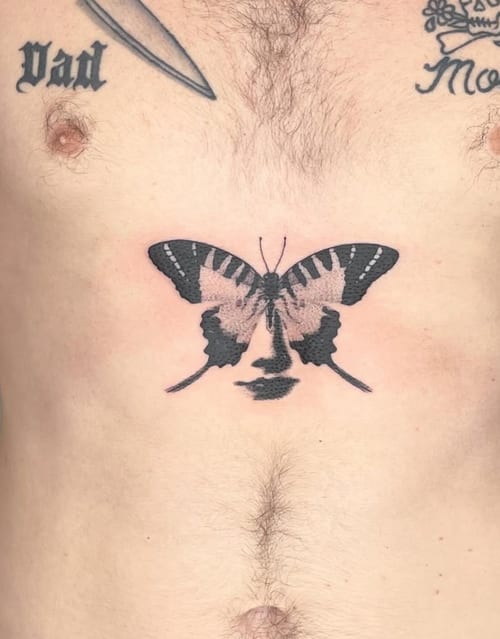 A black and grey surrealistic tattoo of a butterfly with a face integrated into its wings, located on the stomach — by Level tattoo, los angeles
