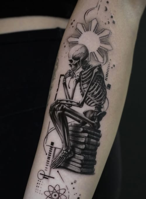 A black and grey tattoo of a skeleton sitting on a stack of books on the forearm, featuring realistic shading and surreal elements — by Jian Liang, los angeles