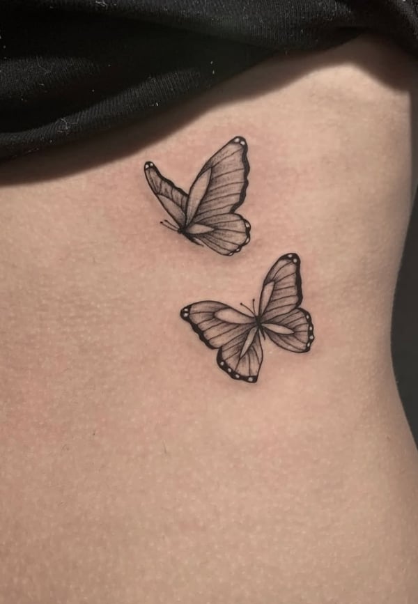 Two black and grey fine line butterflies on the rib, featuring delicate outlines and minimal shading — by Shelby Bianco, ventura, ca