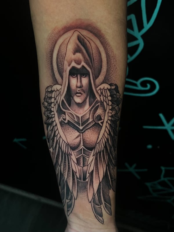 A black and grey realistic depiction of an angel with detailed wings on the forearm, featuring intricate shading and a hooded figure — by Josue Bravo, los angeles