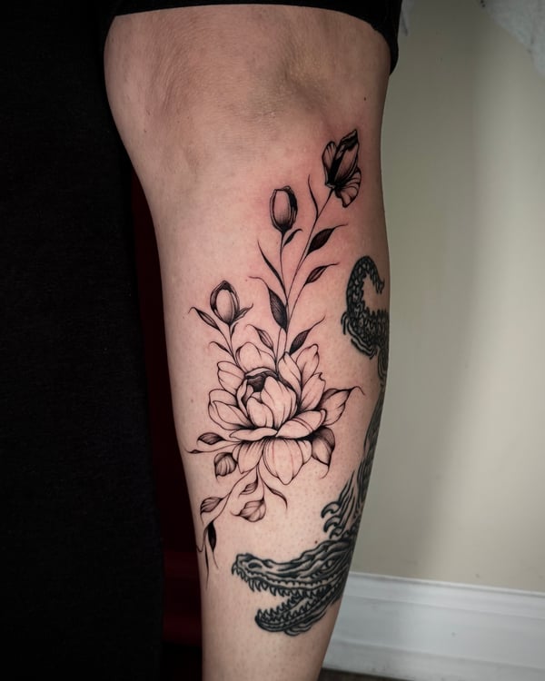 A fine line black and grey floral tattoo on the upper arm, featuring delicate flowers and leaves with intricate linework — by emily, rahway