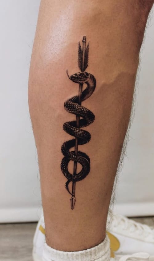 A black and grey tattoo of a snake wrapped around an arrow on the calf, featuring fine line work and detailed shading — by Jian Liang, los angeles