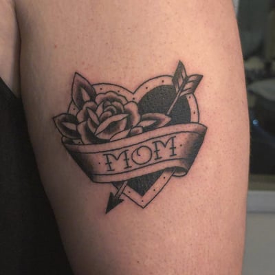 A bold black and grey traditional heart tattoo on the upper arm, featuring a rose and banner with 'Mom' lettering — by Miguel ANGELO, los angeles