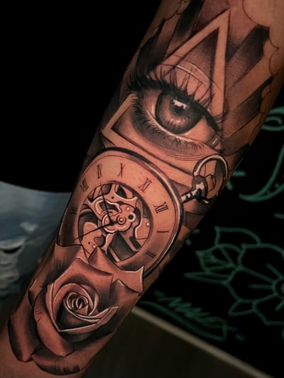 A realistic black and grey tattoo on the forearm featuring a detailed eye, a pocket watch with Roman numerals, and a rose, all intricately shaded — by Josue Bravo, los angeles