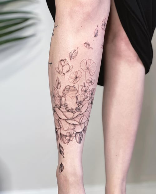 A delicate black and grey fine line tattoo of a frog sitting among flowers on the calf — by Melanie Benoit, los angeles