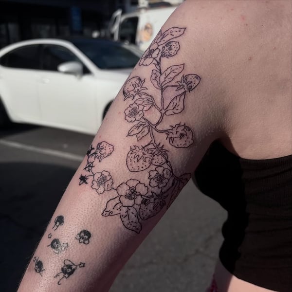 A delicate black linework tattoo of a strawberry plant on the upper arm, featuring flowers and fruit — by Hannah , fountain valley, ca