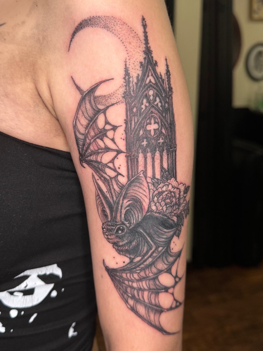 A black and grey tattoo of a bat with gothic architecture and floral elements on the upper arm, featuring dotwork shading and a crescent moon — by Sandra Price, los angeles