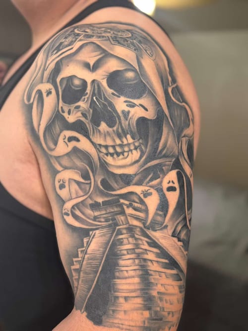 A black and grey tattoo on the upper arm featuring a realistic skull with a hood, surrounded by ghostly figures and a pyramid below — by Eddie Rodriguez, los angeles