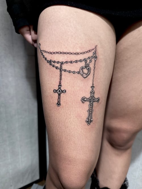 A black and grey fine line tattoo on the thigh featuring two ornate crosses and a heart pendant hanging from chains — by Shelby Bianco, los angeles