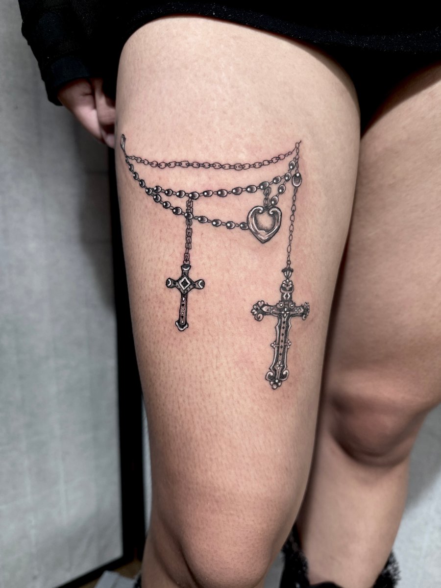 A black and grey fine line tattoo on the thigh featuring two ornate crosses and a heart pendant hanging from chains — by Shelby Bianco, los angeles