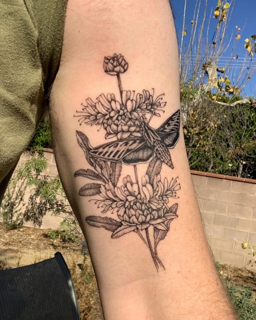 A delicate black and grey tattoo of a moth resting on flowers, located on the upper arm — by Melanie Benoit, los angeles