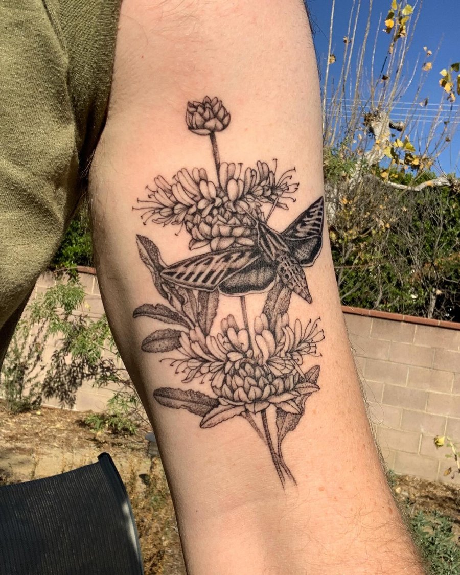 A delicate black and grey tattoo of a moth resting on flowers, located on the upper arm — by Melanie Benoit, los angeles