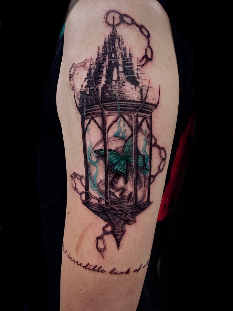 An illustrative tattoo on the upper arm featuring a skull and a butterfly inside a lantern, with chains and a castle in the background. The design uses a mix of black ink and subtle color accents —…