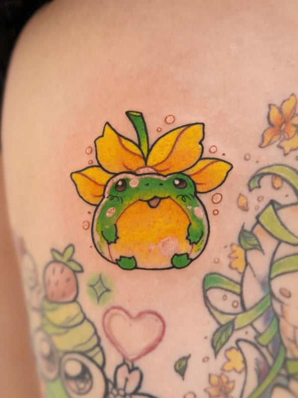 A colorful kawaii-style tattoo of a cute, round animal with a flower on its head, featuring bright greens and yellows on the arm — by Tweetwee, west covina, ca