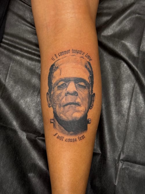 A black and grey portrait of Frankenstein's monster on the calf, surrounded by a quote in bold lettering — by lunayflorestattoo, los angeles