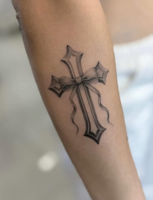 A black and grey fine line tattoo of a cross with a ribbon on the forearm, featuring delicate shading and detail — by otaybeatdown, los angeles