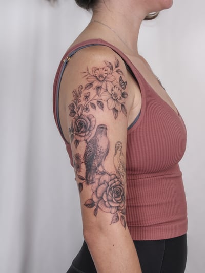 A black and grey tattoo featuring a bird and floral arrangement on the upper arm, showcasing realistic detail and delicate shading — by Melanie Benoit, los angeles