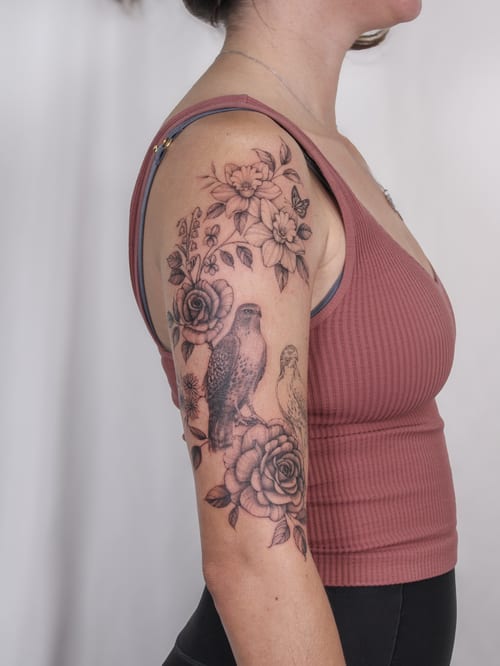 A black and grey tattoo featuring a bird and floral arrangement on the upper arm, showcasing realistic detail and delicate shading — by Melanie Benoit, los angeles