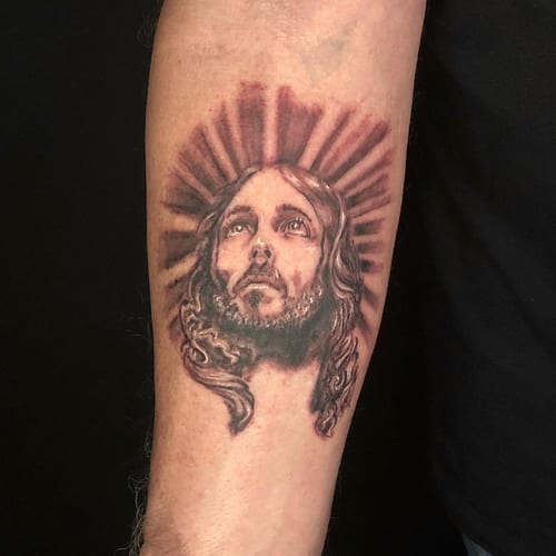 A realistic black and grey portrait of Jesus Christ with a halo on the forearm, showcasing spiritual and ornate details — by Sandra Price, los angeles