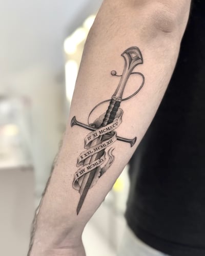A black and grey tattoo of a realistic sword with Roman numerals on the forearm, featuring detailed shading and linework — by otaybeatdown, los angeles