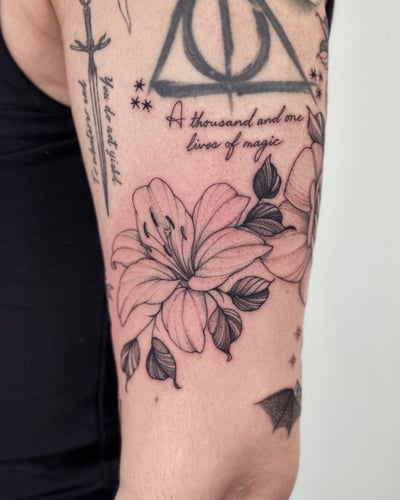 A delicate black and grey lily flower with script on the upper arm, featuring fine line and blackwork style — by Melanie Benoit, los angeles