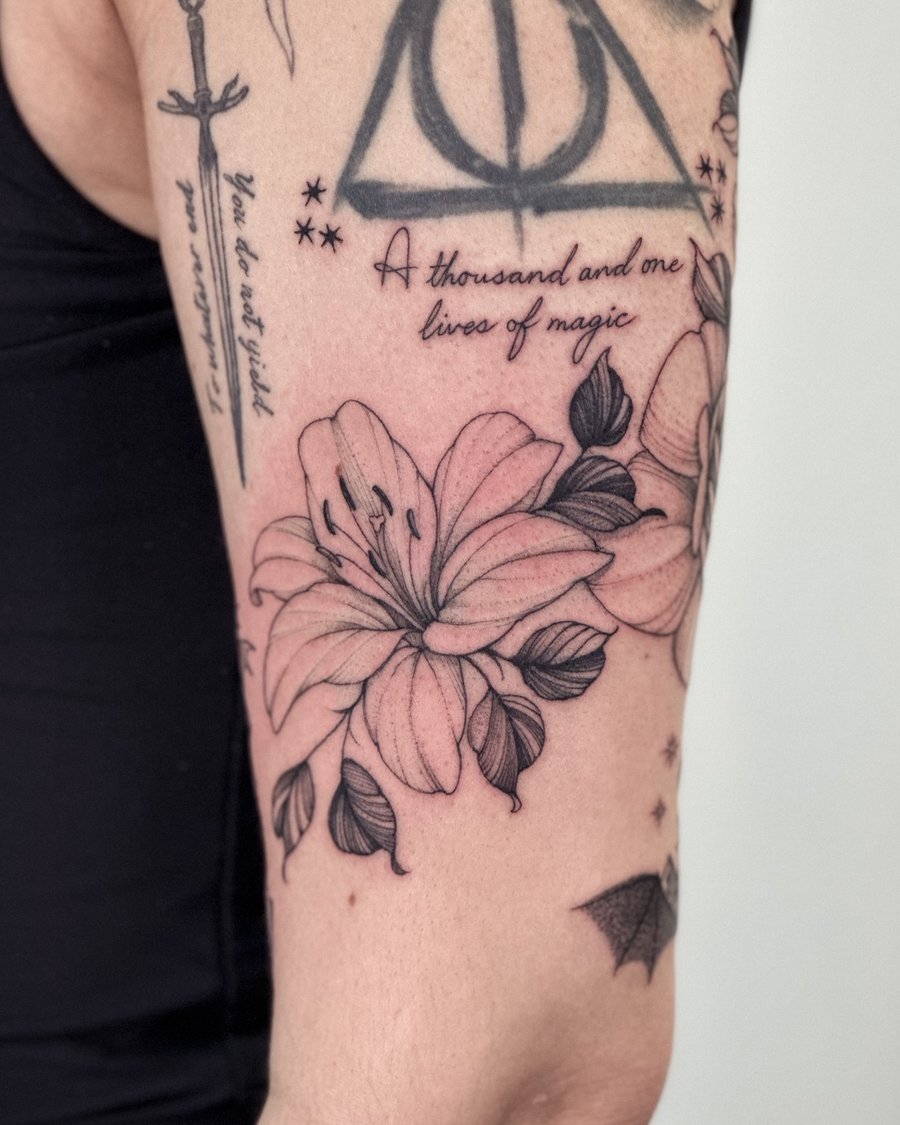 A delicate black and grey lily flower with script on the upper arm, featuring fine line and blackwork style — by Melanie Benoit, los angeles