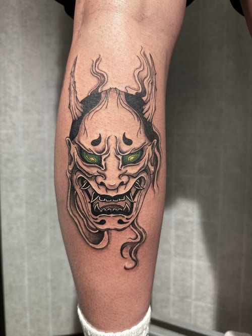 A Japanese-style demon mask tattoo on the calf, featuring bold black outlines and green eyes, with intricate details and shading — by Shelby Bianco, los angeles