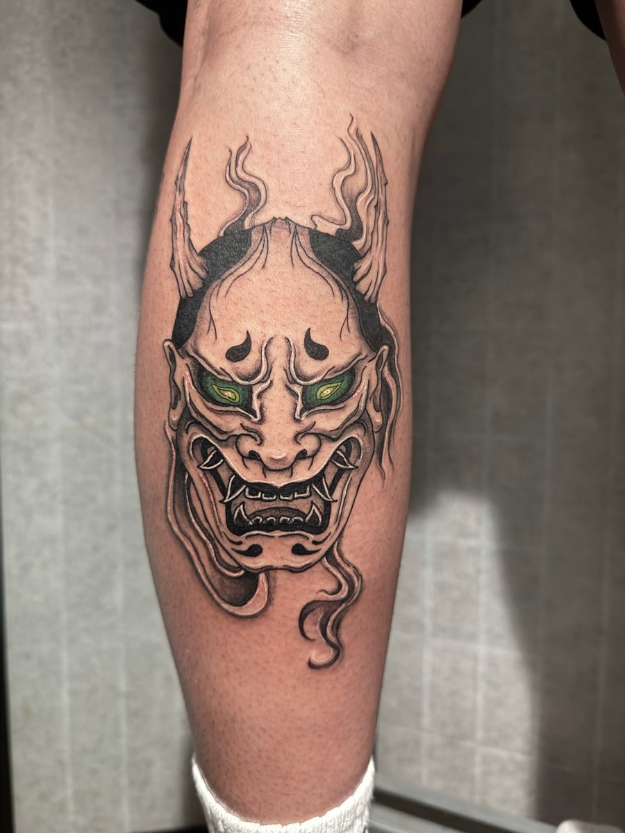 A Japanese-style demon mask tattoo on the calf, featuring bold black outlines and green eyes, with intricate details and shading — by Shelby Bianco, los angeles