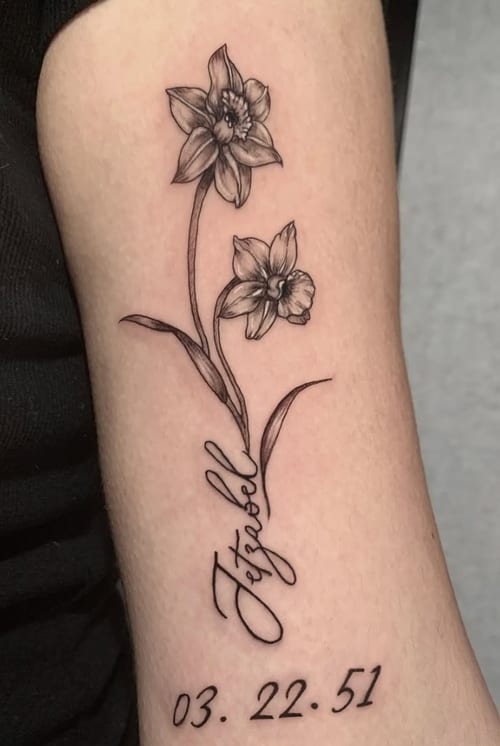 A fine line black and grey tattoo of two flowers with delicate script and a date on the forearm — by Shelby Bianco, los angeles