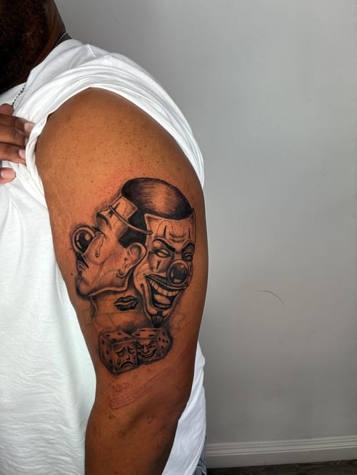 A black and grey Chicano style tattoo on the upper arm featuring two faces with clown-like expressions and a pair of dice, showcasing detailed shading and linework — by Edgar Armas Rodriguez, los a…