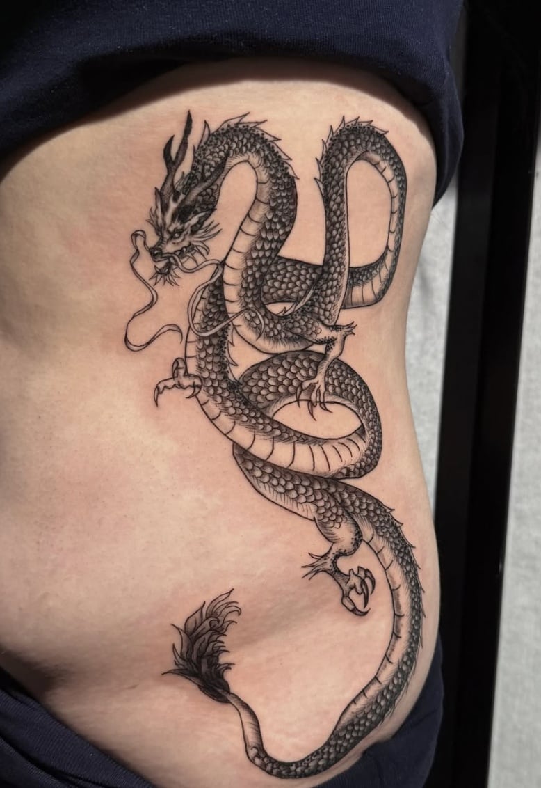 A detailed black and grey dragon tattoo on the rib, featuring intricate scales and flowing lines in a Japanese style — by Shelby Bianco, los angeles