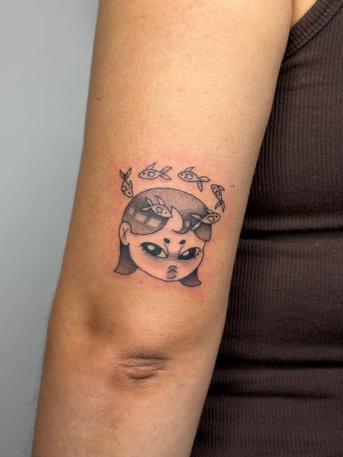 A small, colorful kawaii-style tattoo of a woman's face with fish swimming around her head on the upper arm — by lunayflorestattoo, los angeles
