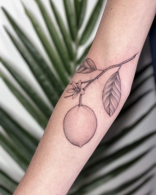 A delicate black and grey fine line tattoo of a lemon branch on the forearm, featuring detailed leaves and a flower — by Melanie Benoit, los angeles