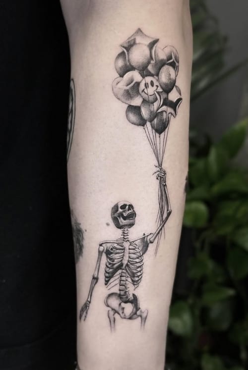 A black and grey illustrative tattoo of a skeleton holding a bunch of balloons on the forearm, with clear linework and shading — by Jian Liang, los angeles