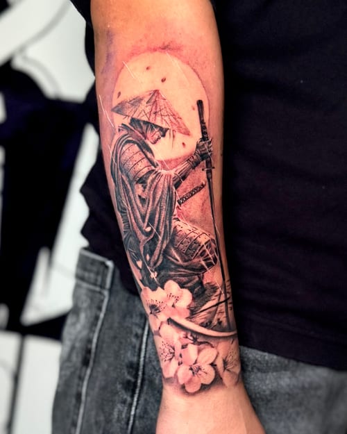 A black and grey realism tattoo of a samurai with a sword, surrounded by cherry blossoms, on the forearm — by zealous, los angeles