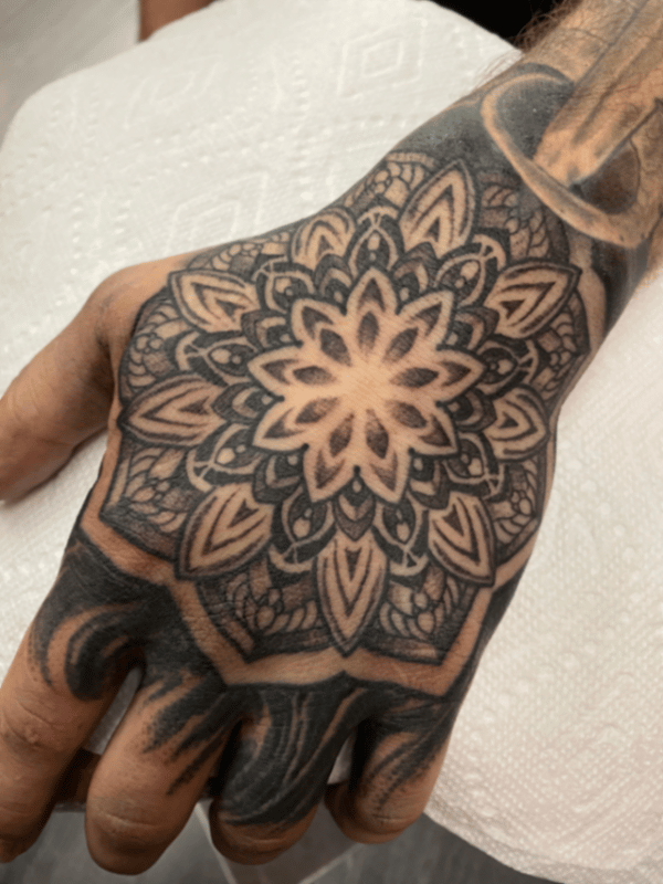 A detailed black and grey mandala tattoo covering the hand with intricate patterns and bold lines — by tyler_whitlock, los angeles