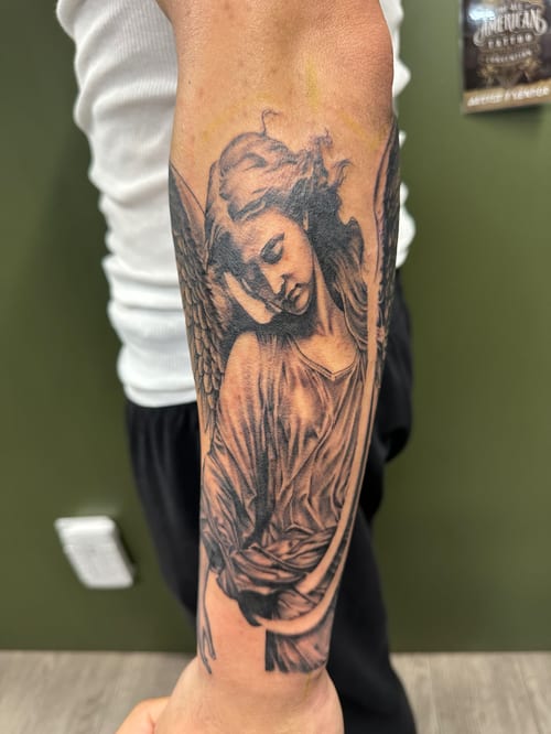 A detailed black and grey realism tattoo of an angel on the forearm, featuring intricate shading and soft textures — by Humilde tattoos, los angeles