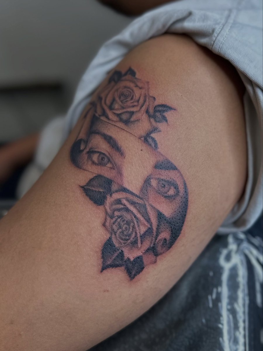 A bold black and grey tattoo of a woman's eyes surrounded by roses on the upper arm — by Maddie Ramirez, los angeles