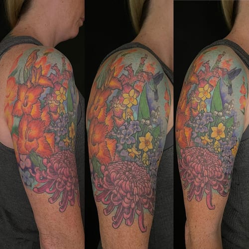 A vibrant color tattoo of a floral arrangement with a hummingbird on the upper arm, featuring detailed flowers and leaves — by Sandra Price, los angeles
