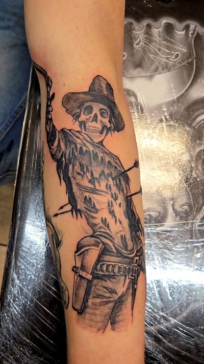 A black and grey tattoo of a skeleton wearing a hat and tattered clothing on the forearm, featuring bold lines and dark shading — by Dretty, los angeles