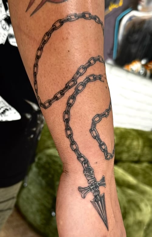 A black and grey tattoo of a chain wrapped around the forearm ending in a dagger, featuring bold lines and detailed shading — by otaybeatdown, los angeles