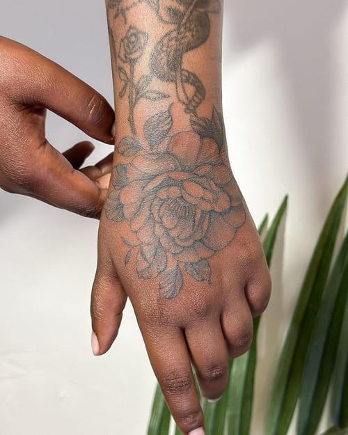 A delicate black and grey floral tattoo on the hand, featuring a large flower with detailed petals — by Melanie Benoit, los angeles
