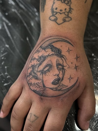 A delicate fine line tattoo of a crying moon face on the hand, done in black and grey — by Maddie Ramirez, los angeles
