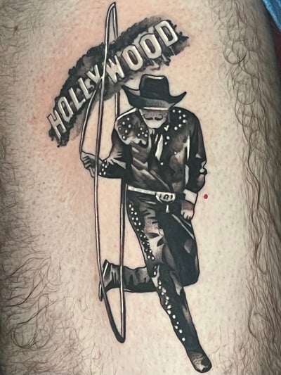A black and grey illustrative tattoo on the leg depicting a cowboy figure with the word 'Hollywood' above, rendered in a bold blackwork style — by Josue Bravo, los angeles