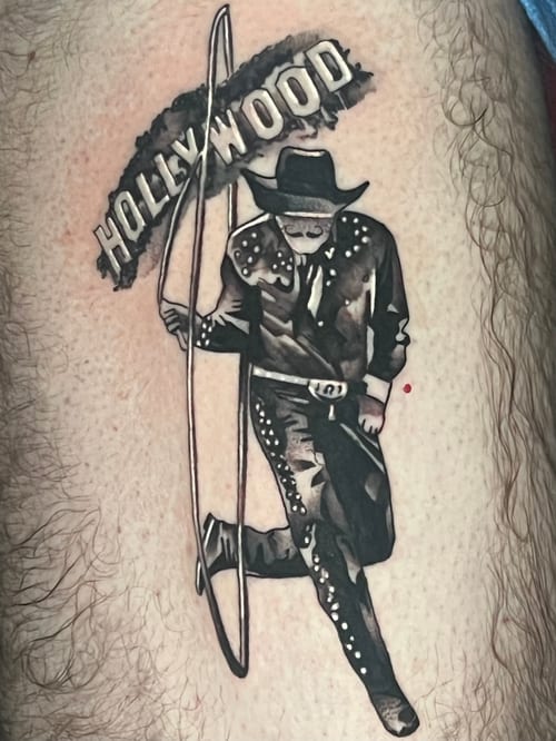 A black and grey illustrative tattoo on the leg depicting a cowboy figure with the word 'Hollywood' above, rendered in a bold blackwork style — by Josue Bravo, los angeles