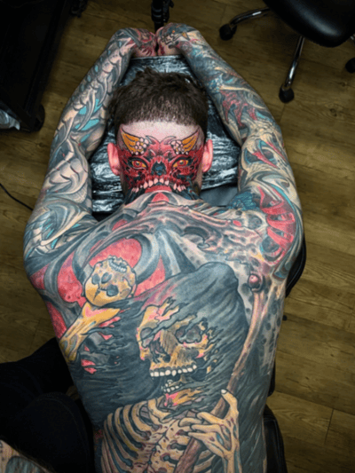 A colorful neo-traditional tattoo covering the full back, featuring a skeleton holding a skull with a demonic face on the neck. The design includes vivid colors and intricate details — by tyler_whi…