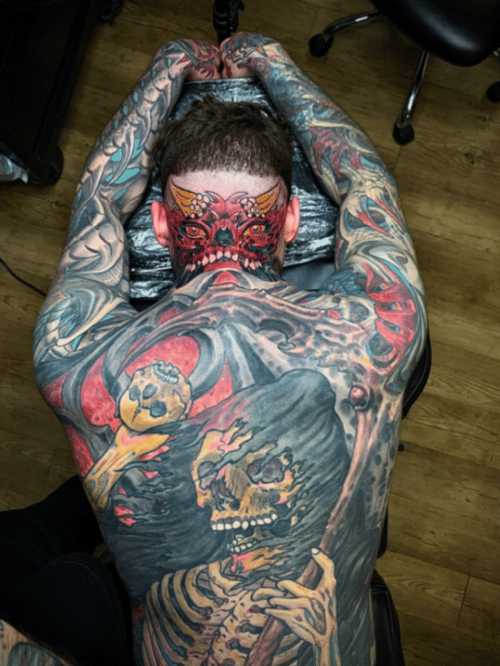 A colorful neo-traditional tattoo covering the full back, featuring a skeleton holding a skull with a demonic face on the neck. The design includes vivid colors and intricate details — by tyler_whi…